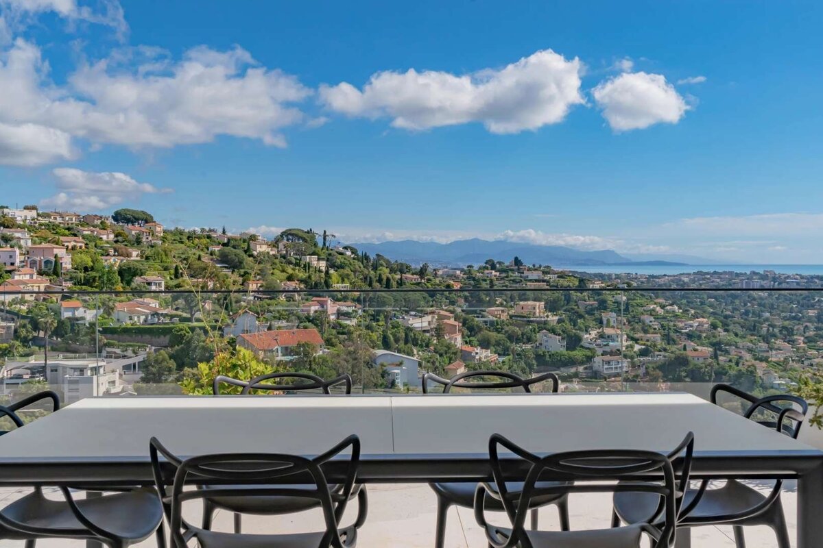A grey outdoor dining table with black chairs frames a panoramic view of a green, hilly town, the distant sea, and mountains under a blue, cloudy sky.