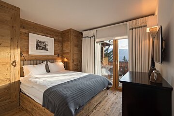 Cozy wood-paneled bedroom with a large bed, rustic decor, and glass doors opening to a scenic mountain balcony view.