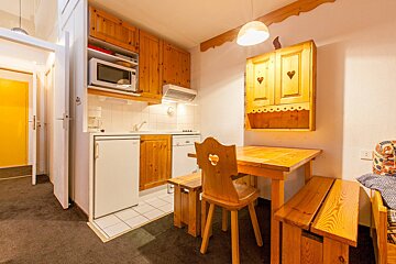 A compact, rustic kitchen and dining area in a chalet style, featuring light wooden cabinets, a table with heart-cutout chairs, a fridge, and a microwave.
