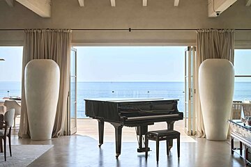 A black grand piano in a luxurious room with large open doors leading to a vast ocean view, flanked by two tall white vases.