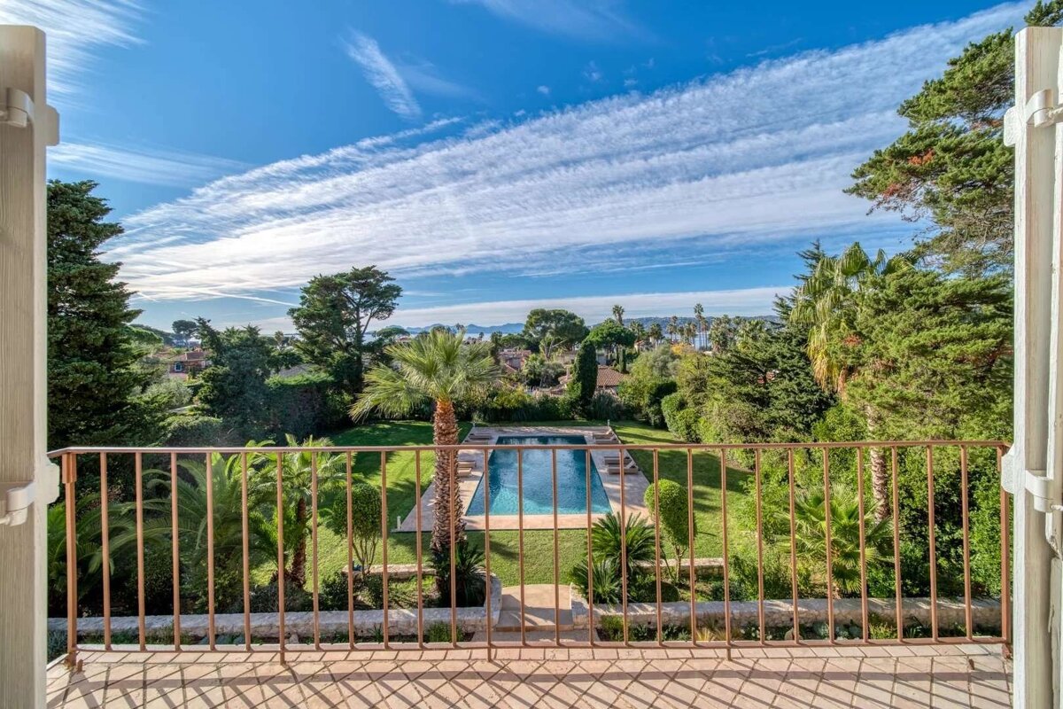 A balcony view overlooks a lush garden with a swimming pool, palm trees, and distant sea under a bright blue sky with streaky clouds.