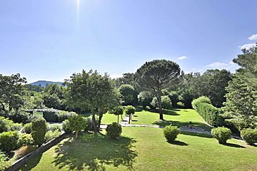 A bright, sunny view of a large green garden with diverse trees, bushes, and pathways. Mountains are visible in the background under a clear blue sky.