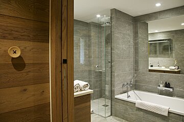 A modern bathroom features grey tiles, a walk-in glass shower, and a bathtub. Warm wooden accents, including a unique button-shaped wall hook, add character.