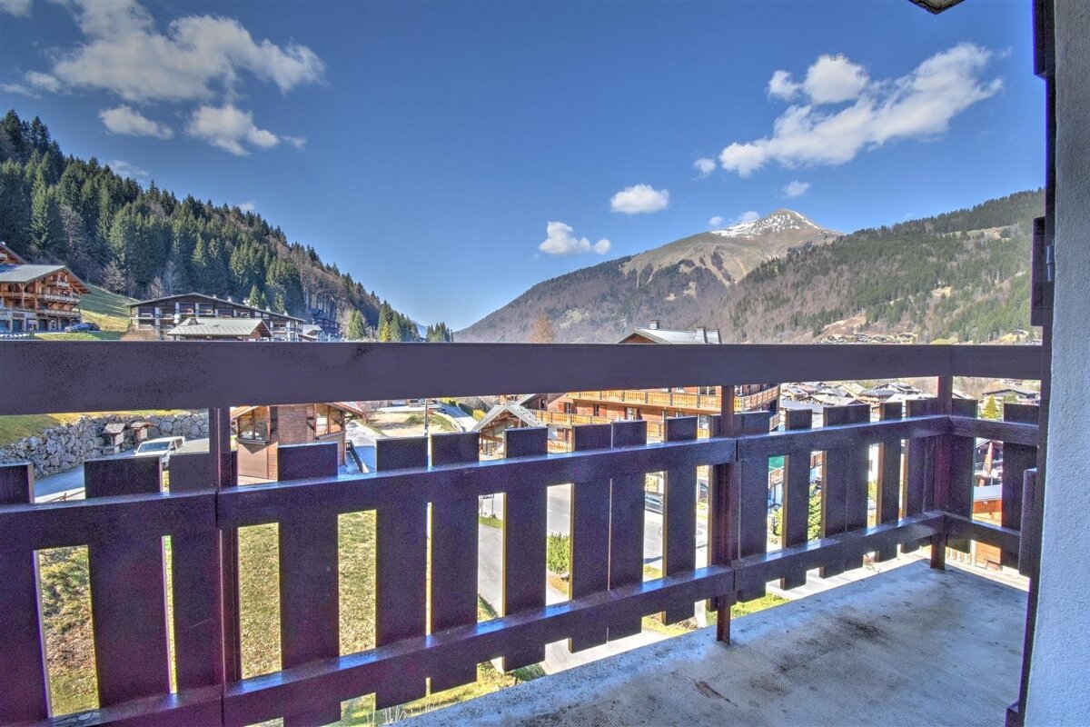 Scenic view of an alpine village and mountains under a bright blue sky, seen from a wooden balcony.