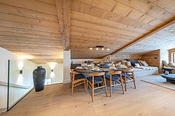 A modern chalet-style attic room with extensive light wood paneling, a dining table set, and a cozy living area. Features a glass railing.
