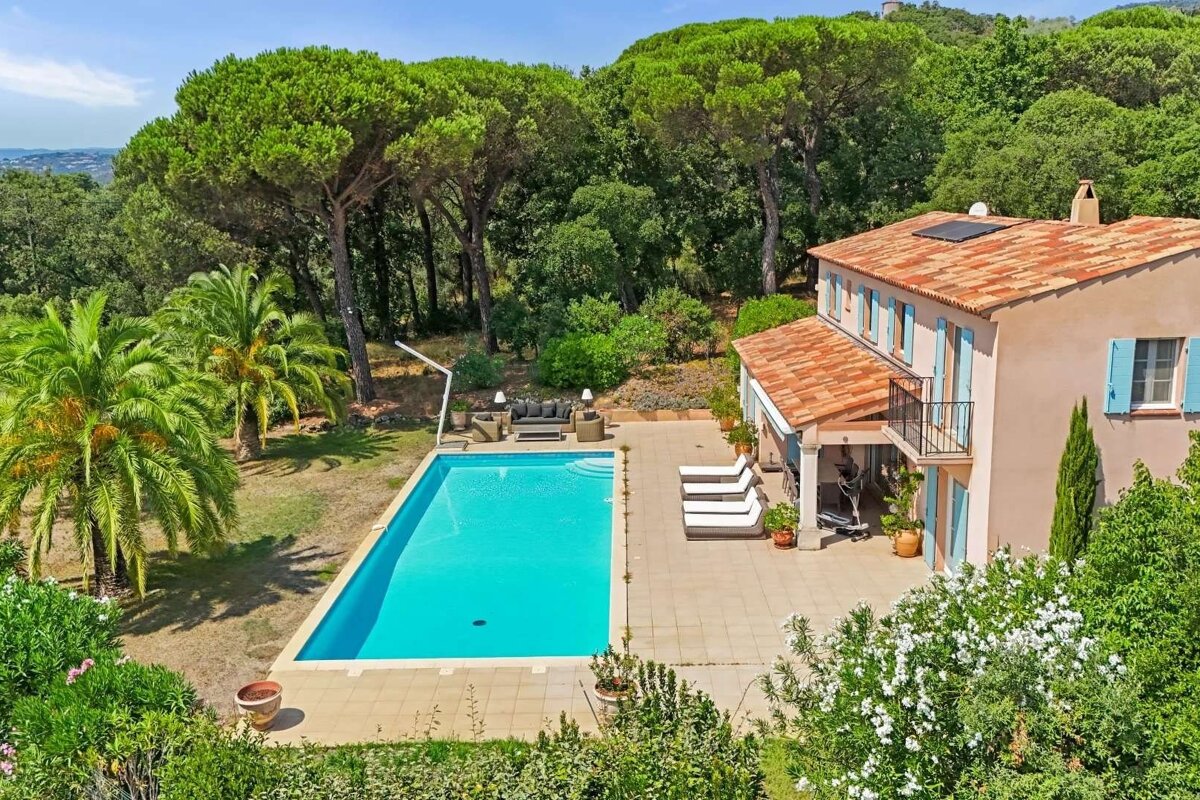 Aerial view of a house with red roof and blue shutters, a sparkling pool, and patio with loungers, all nestled among lush green trees and foliage.