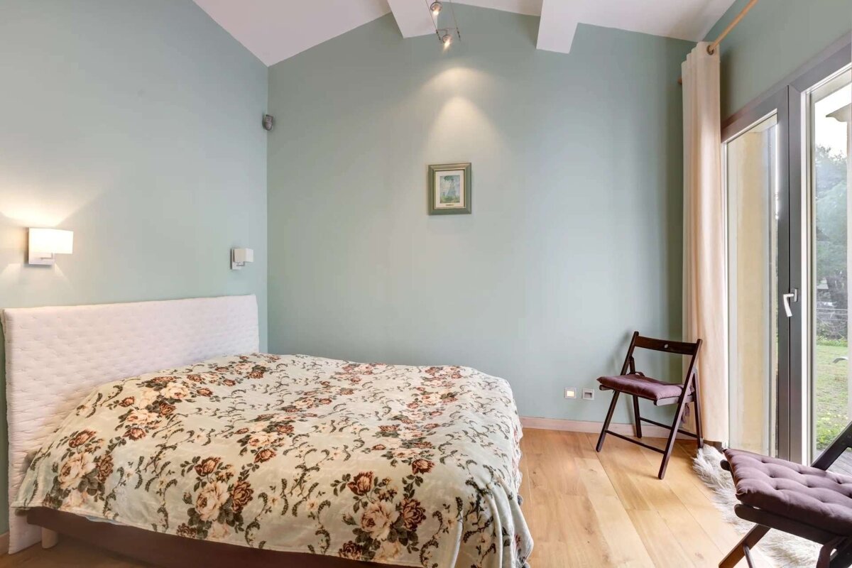 A bedroom with pale green walls, a floral bedspread, hardwood floors, and high ceilings. Features include wall lights, a small framed picture, and a glass door.