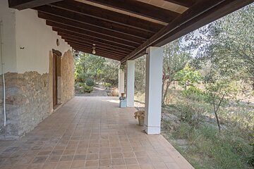 A covered outdoor patio with tiled floor, white columns, and a wooden ceiling, alongside a stone wall, facing a rustic garden with trees.