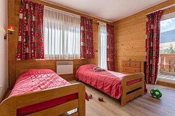 A cozy, wood-paneled room features two single beds with red bedding, festive red curtains, and a balcony door overlooking mountains.