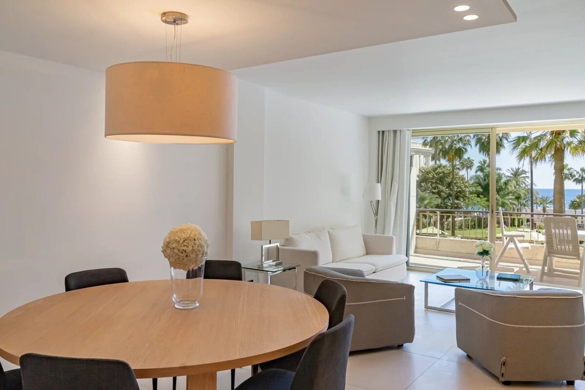 A modern living and dining area in a bright apartment, featuring neutral tones and a balcony view of palm trees and the ocean.
