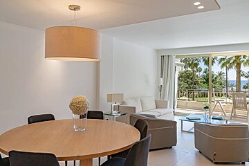 A bright, modern dining and living room in neutral tones, featuring a large window opening to a balcony with palm trees.