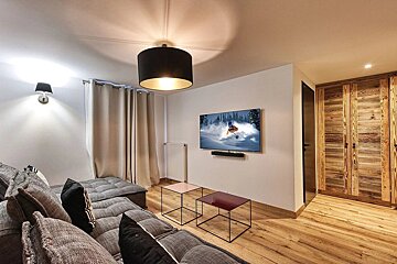 A cozy, modern living room with a large grey sectional sofa, a TV displaying skiing, wooden flooring, and rustic wooden paneling.