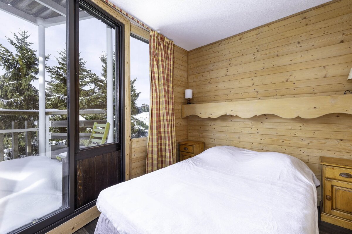 Cozy wood-paneled bedroom with a white bed, featuring a large glass door and window leading to a snowy balcony with pine trees.