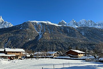 Snowy mountain village with wooden chalets, pine forests, and towering jagged peaks under a clear blue sky.