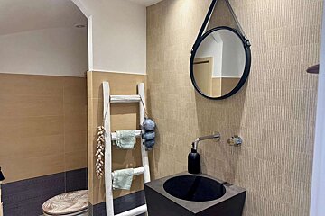 A modern bathroom with a black sink, round mirror, and decorative ladder holding towels. Earthy tiled walls and a palm-leaf toilet lid create a natural vibe.