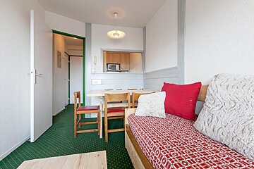 A small apartment living room with green patterned carpet, a red sofa bed, a dining table with wooden chairs, and a kitchen visible through a pass-through.