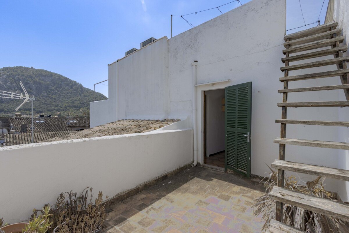 A sunlit rooftop terrace features a tiled floor, white walls, an open green door, and wooden stairs, with a lush mountain visible under a clear blue sky.