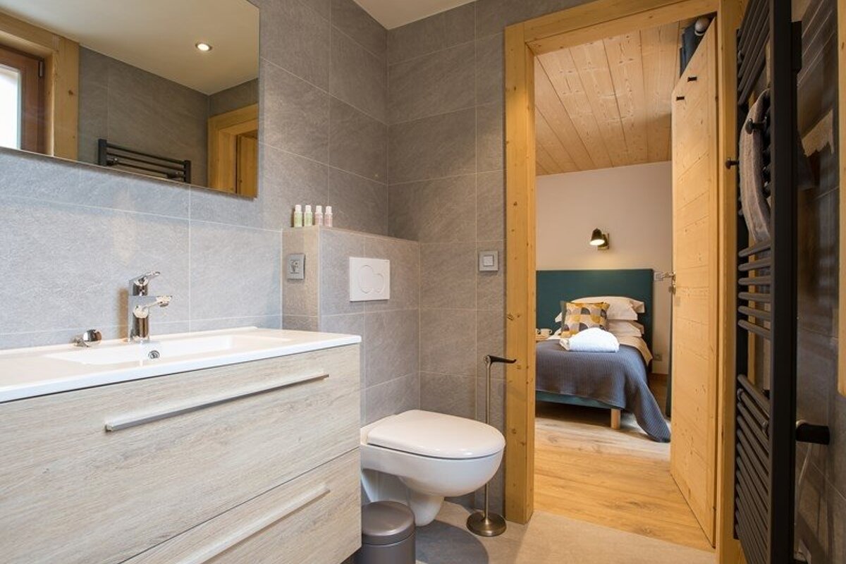 Modern grey-tiled bathroom with a wooden vanity looking through an open doorway into a cozy bedroom.