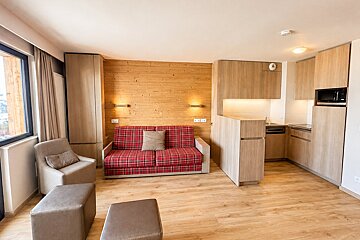 A modern studio apartment featuring a red plaid sofa, light wood paneling, an armchair, and a small kitchenette.