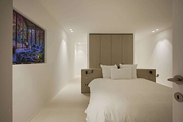 A modern, minimalist bedroom featuring a large white bed, built-in wardrobe, and a unique, trapezoidal screen displaying a cozy autumn living room scene.