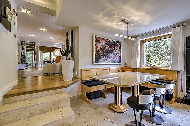 A modern split-level room: a tiled dining area with a long bench and marble table leads up to a wooden living room. A large window views trees.