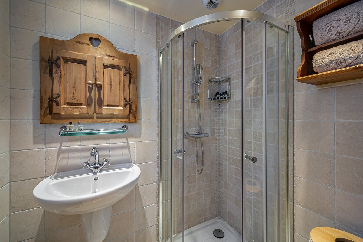 A rustic tiled bathroom featuring a pedestal sink, a corner glass shower, a wooden cabinet with a heart, and a shelf holding towels.