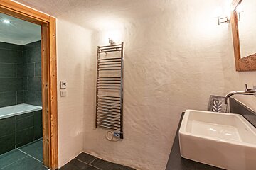 A modern bathroom with a white basin, heated towel rail, and an adjacent dark-tiled room featuring a bathtub.