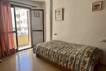 Simple bedroom with single bed, patterned quilt, and open balcony to urban view. Light walls display art.