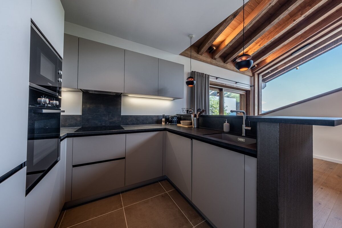 Sleek modern kitchen with gray cabinets, dark counters, integrated appliances, wooden ceiling beams, and a large window view.