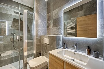A modern bathroom features grey tiled walls, a glass shower, toilet, and a vanity with a white sink and light wood cabinet. A backlit mirror illuminates the space.