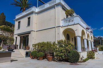 A stately cream-colored villa featuring white balustrades, arched columns, and green landscaping. A palm tree tops the roof under a clear blue sky.