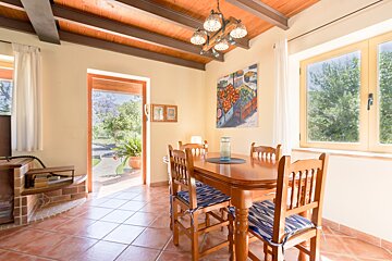 An inviting dining room with a wooden table, terracotta floor, exposed beams, and a colorful fruit painting. It offers bright views of a lush garden.