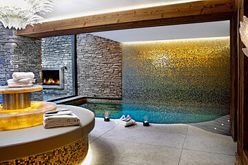 Luxurious spa room with a gold-to-blue mosaic pool wall, stone accents, a cozy fireplace, and soft lighting, offering a serene escape.