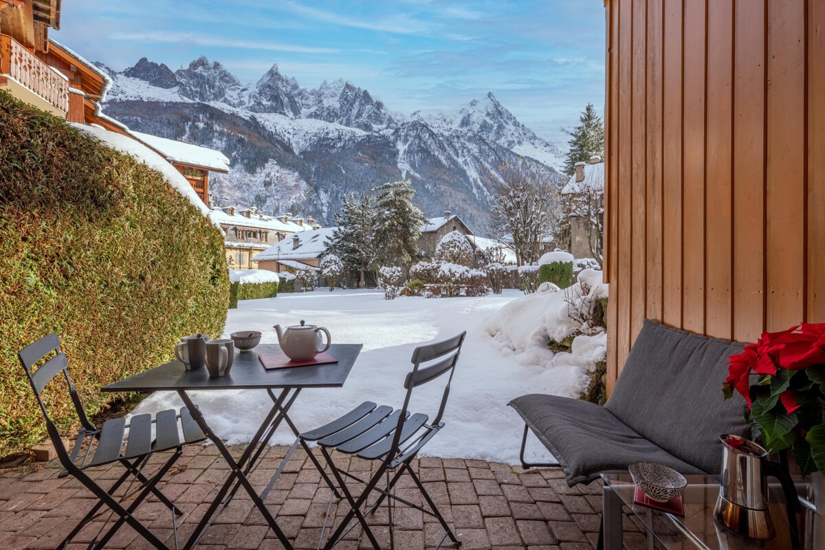 A cozy winter patio with a tea set overlooks a snowy village and majestic mountains under a bright blue sky.