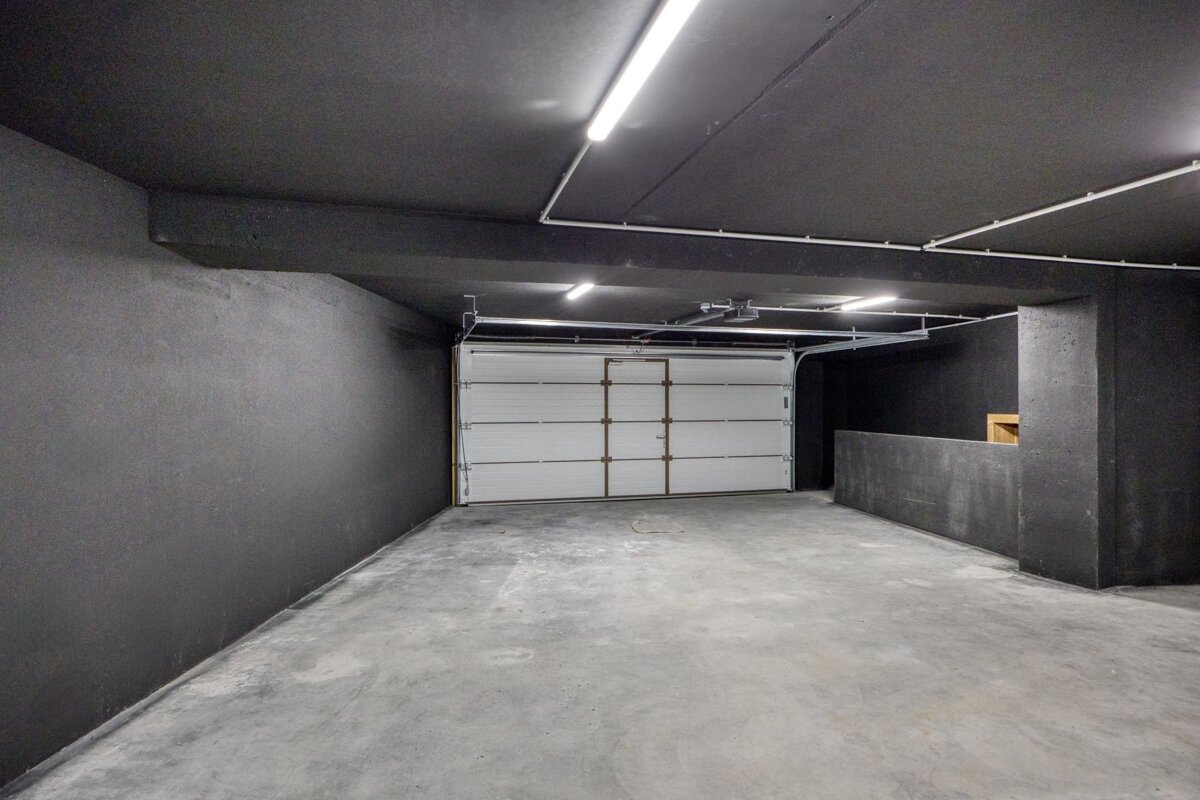 An empty garage with dark grey walls, a concrete floor, and a white garage door lit by overhead tube lights.