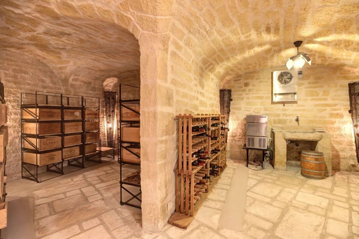 An old-world stone wine cellar with arched ceiling, filled with wine bottles, wooden cases, a stone basin, and a wooden barrel.