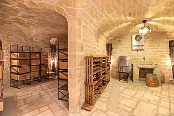 A charming vaulted stone wine cellar with racks of wine bottles, wooden crates, a rustic stone sink, and a wooden barrel on a stone floor.