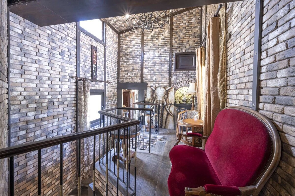 A multi-level, rustic room with exposed brick walls, a winding staircase, red velvet armchair, elephant art, and a chandelier, creating an opulent feel.