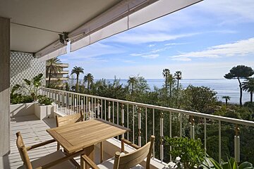 A sunny balcony with wooden furniture overlooks a blue ocean, palm trees, and lush greenery under a clear sky.