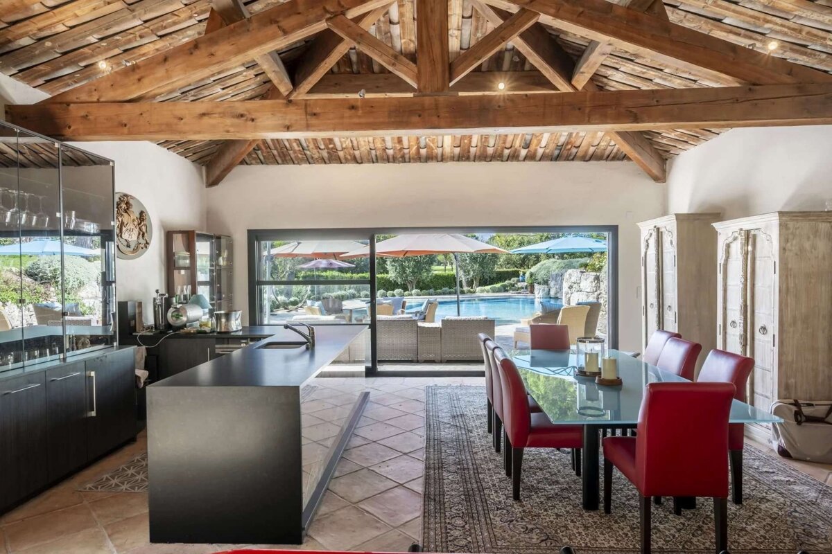 Stylish open-plan kitchen and dining area with exposed wood beams, modern black counters, red chairs, and large windows revealing a pool and patio.