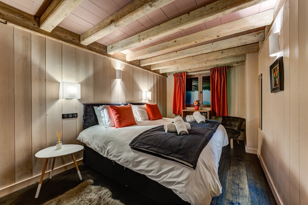 A warm, rustic bedroom with a large bed, orange accents, wooden paneled walls, and exposed ceiling beams. Red curtains frame a window, with soft lighting.