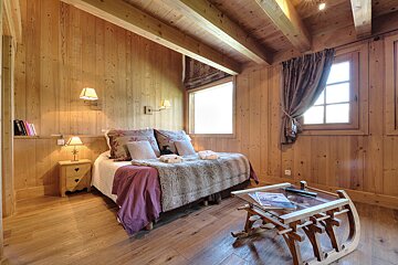 A cozy, rustic chalet bedroom with extensive wood paneling, a double bed, and a unique sled-shaped coffee table by two bright windows.