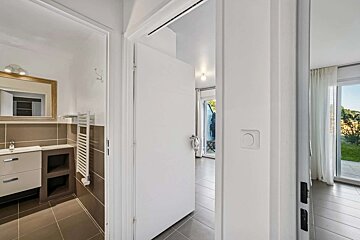 An open white door in a bright interior. To the left is a modern bathroom with a vanity, and to the right, a room with a balcony view.