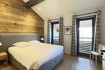 A rustic-modern bedroom with a large bed against a wood-paneled wall, wooden floor, and two balcony doors with plaid curtains.