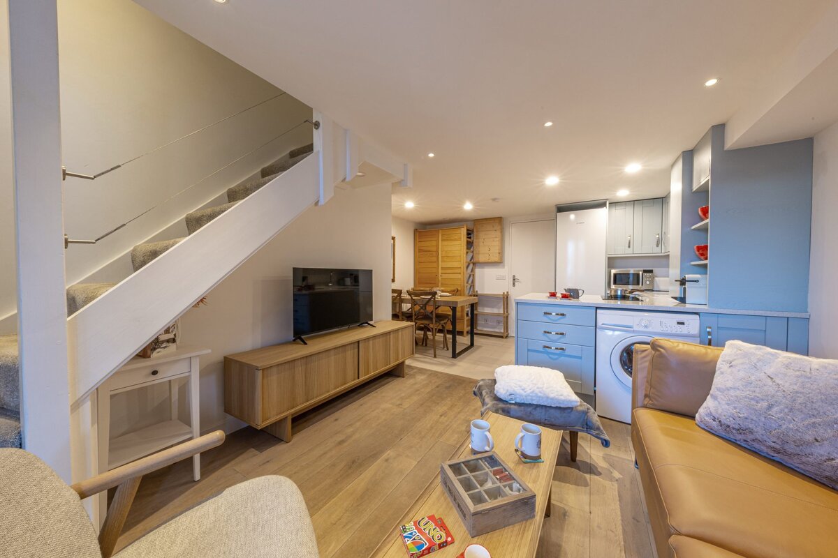 A modern open-plan living room with light blue kitchen, wooden furniture, brown sofa, and white carpeted stairs.