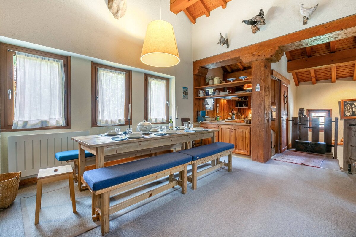 Cozy, rustic dining area with a long wooden table, blue benches, and an open kitchen. Exposed wood beams, high ceiling, and charming decor create a warm ambiance.