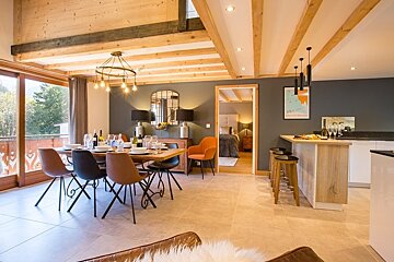 Cozy modern chalet interior with a dining area, kitchen bar, exposed wooden ceiling beams, and warm lighting.