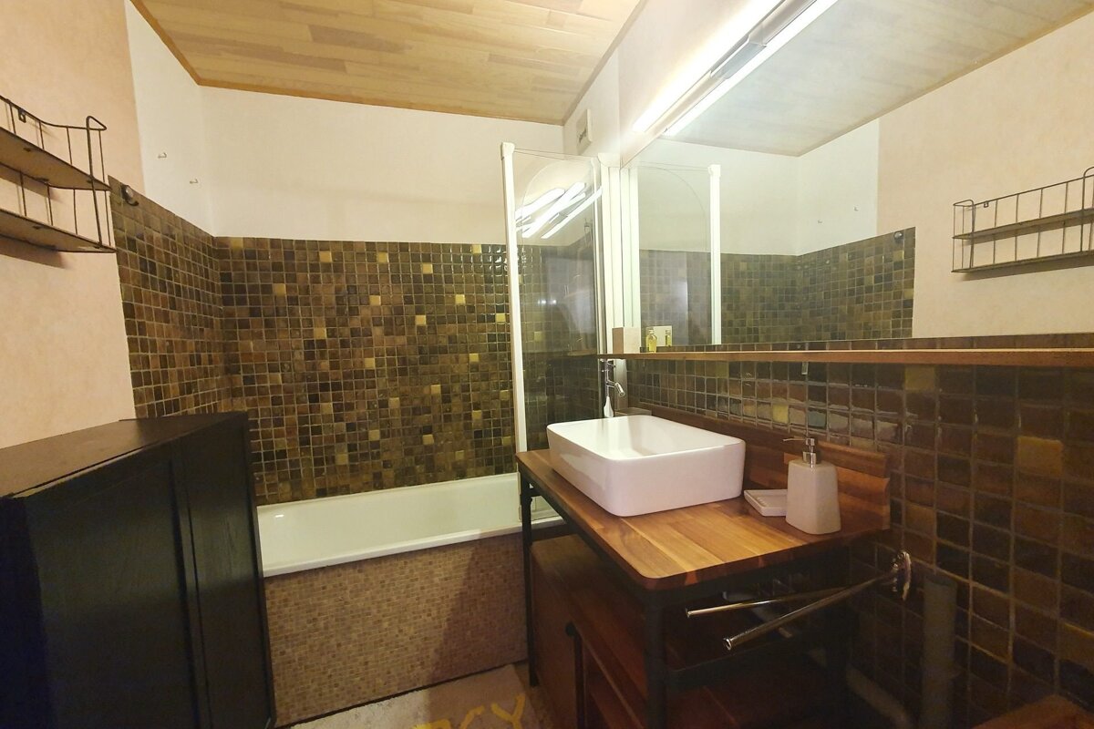 A cozy bathroom with dark mosaic tiled walls, a white bathtub with a shower screen, and a wooden vanity featuring a white vessel sink. A large mirror and wooden ceiling complete the space.