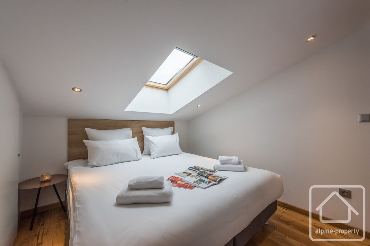 A bright bedroom with a bed featuring white linen, a wooden headboard, and a skylight on the slanted ceiling. Magazines and towels are on the bed.