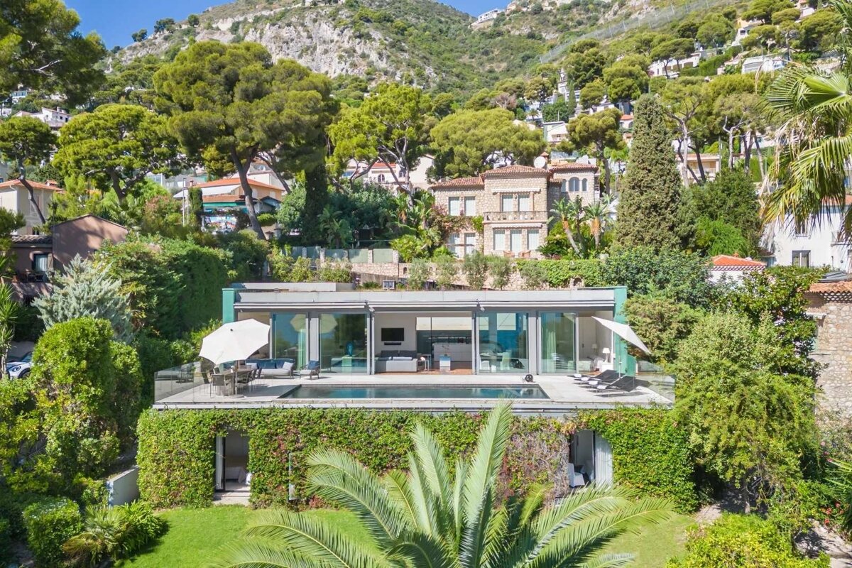 A modern cliffside home with an infinity pool and glass walls, surrounded by lush green foliage and traditional houses, against a mountainous backdrop.